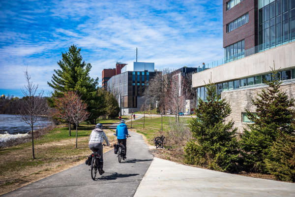 Two people riding their bikes by the Rideau River and the campus. buildings.