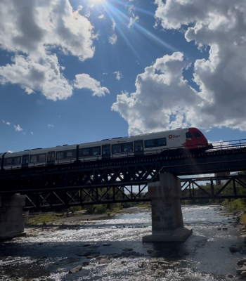 The O-train crosses the Rideau River bridge against a bright blue sky.