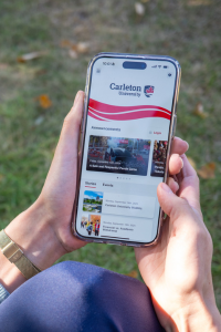 Hands hold an iPhone displaying the Carleton Mobile app home screen.