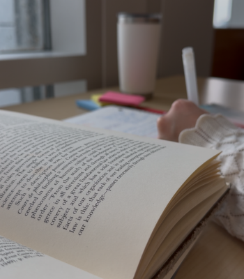 A student hand writes notes in the library with open books, post it notes and a reusable mug.