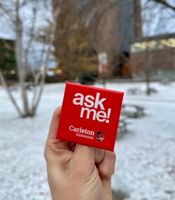 Close-up of 'ask me!' button being held in a snowy quad.