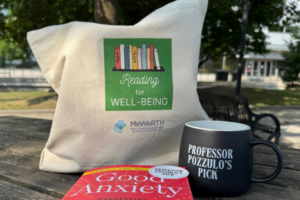 A tote bag and mug branded with the Reading for Well-Being book club, and a paperback with a red cover on a picnic bench.