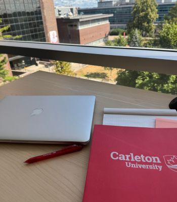 A laptop and a Carleton University notebook on a desk in the MacOdrum library.