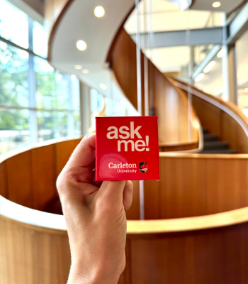 A hand holding a red Ask Me pin in front of the MacOdrum Library staircase.