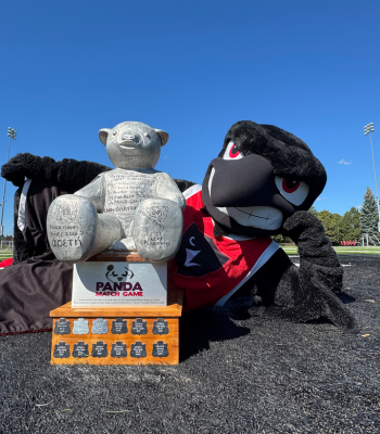 Rodney the Raven mascot lying on the ground with the Panda game award (Pedro) by his side.